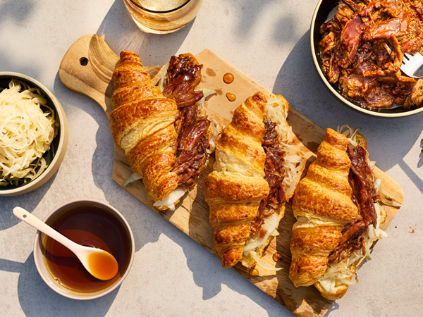 Auf einem Tisch steht ein Holzbrett mit drei Ahorn-Pulled Pork im Croissant. Links steht eine Schüssel mit Weißkraut, dadrunter eine kleine Schüssel mit Ahornsirup mit einem Löffel drin. Rechts steht eine Schüssel mit Ahorn-Pulled Pork.