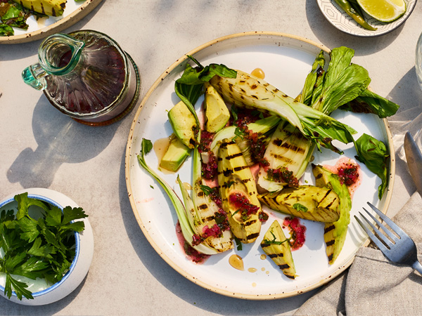 Gegrillter Pak Choi mit Ahornsirup-Limetten-Relish auf einem Teller. Daneben stehen eine Karaffe mit Ahornsirup, frische Kräuter und Limettenspalten.