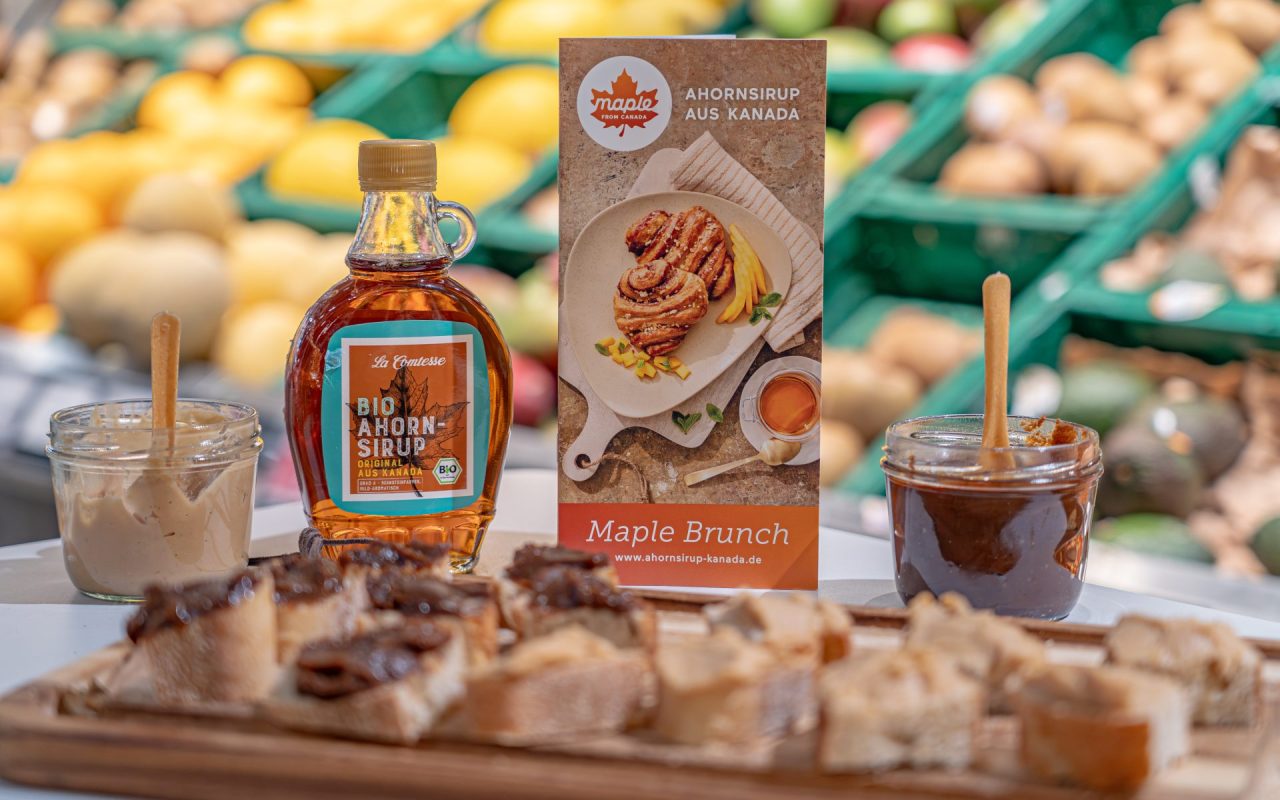 Flasche Ahornsirup aus Kanada, Broschüre "Maple Brunch" und ein Glas Ahorn-Nuss Nougat Creme. Im Vordergrund: Brotscheiben mit Aufstrich.
